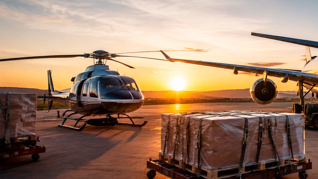 Helicóptero no pátio ao pôr do sol, ao lado de paletes de carga embalados e da asa de um jato