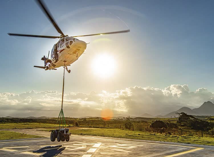 Helicóptero bimotor decolando de heliponto com um trator suspenso por cabo, contra o sol