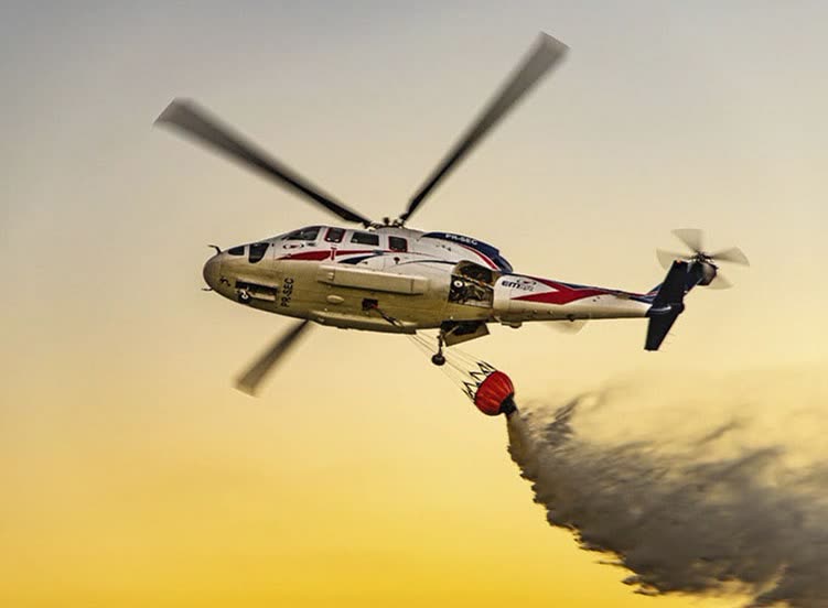 Helicóptero com balde de água (bambi bucket) em combate a incêndio, contra o céu alaranjado