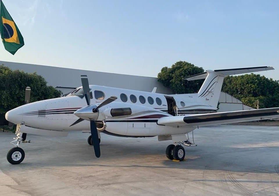 Avião bimotor turboélice branco diante de hangar, com a bandeira do Brasil hasteada ao lado
