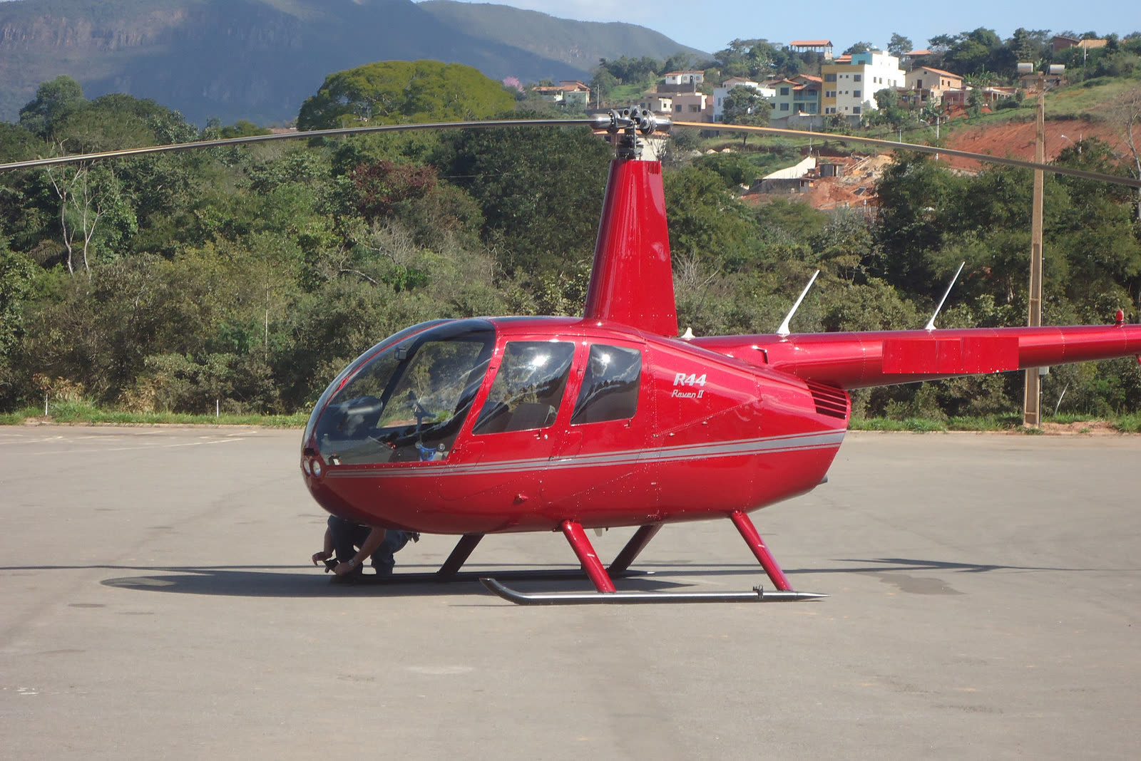 Helicóptero monomotor vermelho pousado em pátio asfaltado, com morros e casas ao fundo