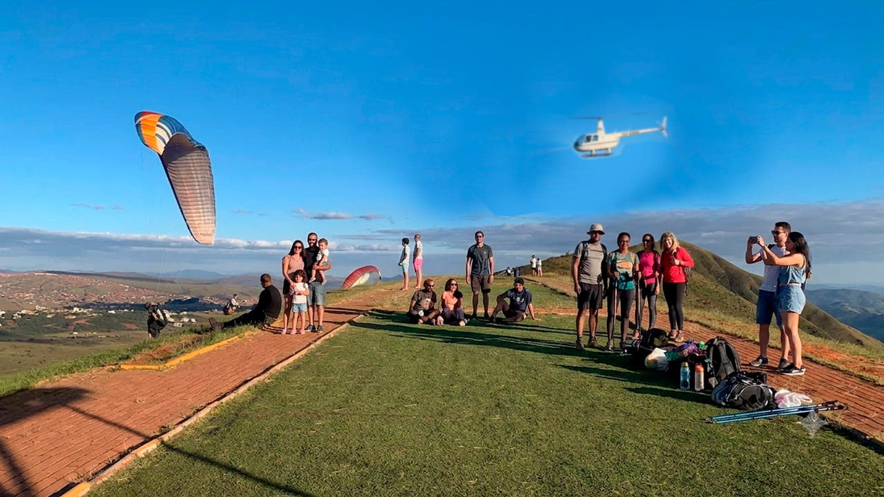 Grupo na rampa de voo livre no alto da serra observa parapente e helicóptero no céu azul