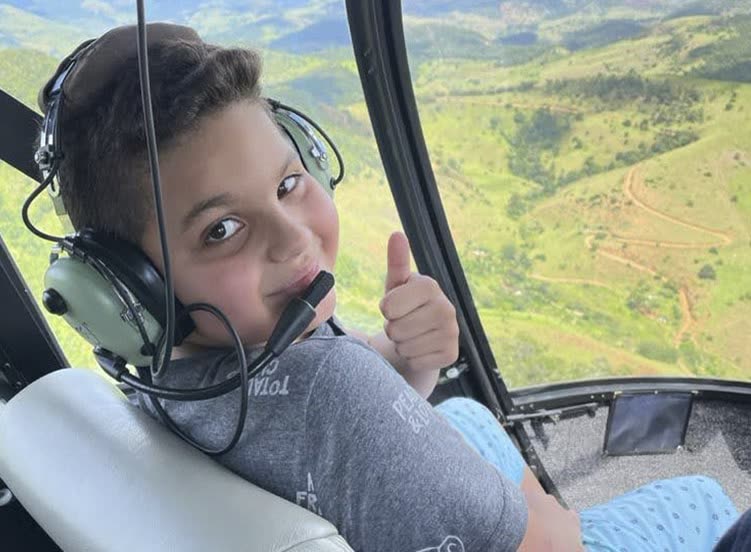 Menino com fone de aviação faz sinal de positivo dentro do helicóptero, em voo sobre morros verdes