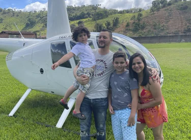 Família sorridente ao lado de helicóptero branco pousado em gramado cercado de morros