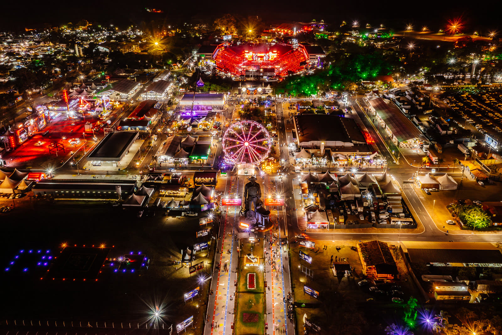 Parque de exposições iluminado à noite visto do alto, com roda-gigante, estátua e avenida central