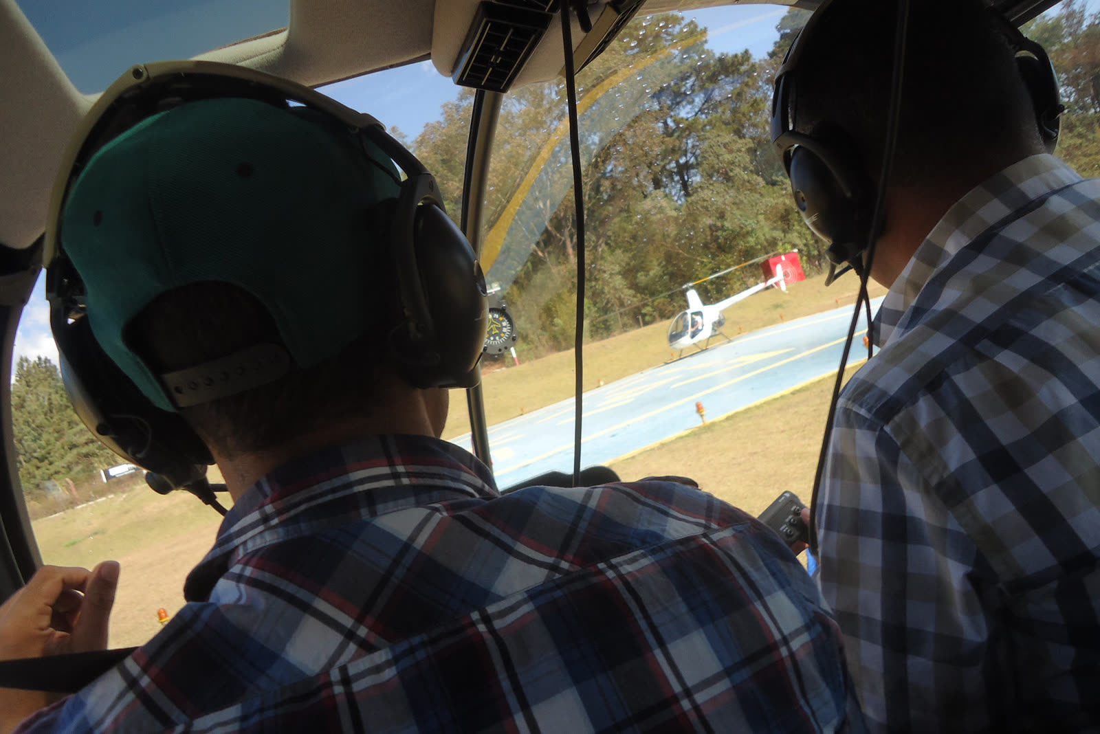 Piloto e passageiro com fones na cabine, com outro helicóptero pousado no heliponto à frente