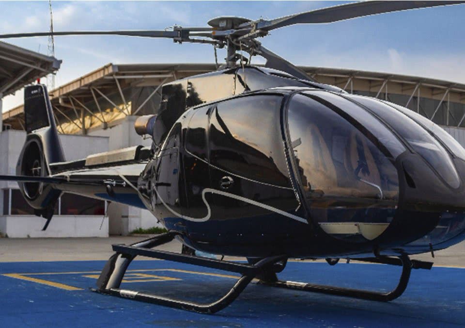 Helicóptero monomotor preto em heliponto azul diante de hangar com cobertura metálica