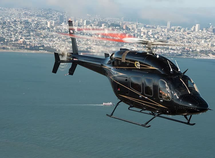 Helicóptero bimotor preto com detalhes dourados voando sobre uma baía, com a cidade ao fundo