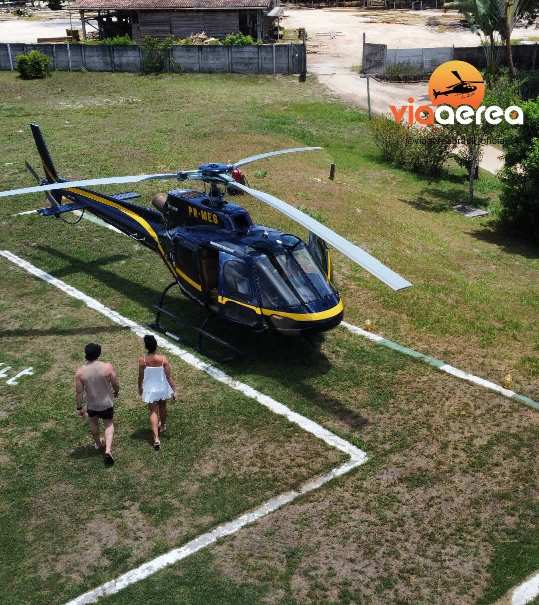 Casal caminhando até helicóptero azul e amarelo pousado em heliponto gramado